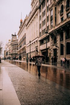 A pedestrian with an umbrella strolls along historic Gran Vía in Madrid on a rainy day.