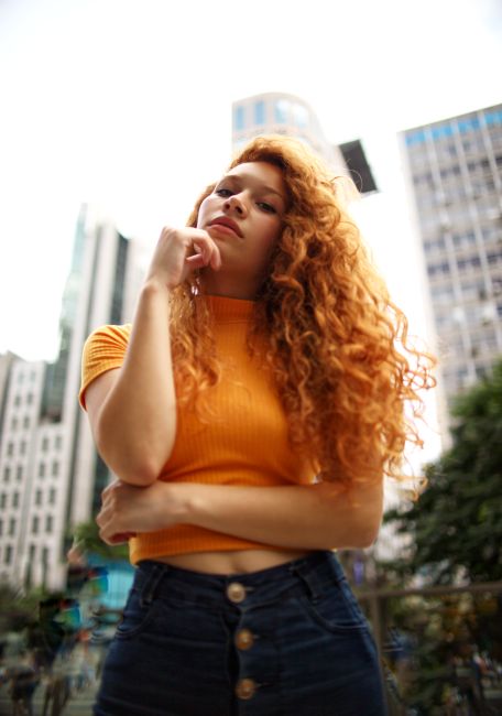 A fashionable young woman with curly red hair poses confidently against a city backdrop.