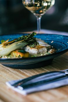 Close-up of a gourmet fish dish served on a blue plate, garnished with roasted pak choi, in a restaurant setting.