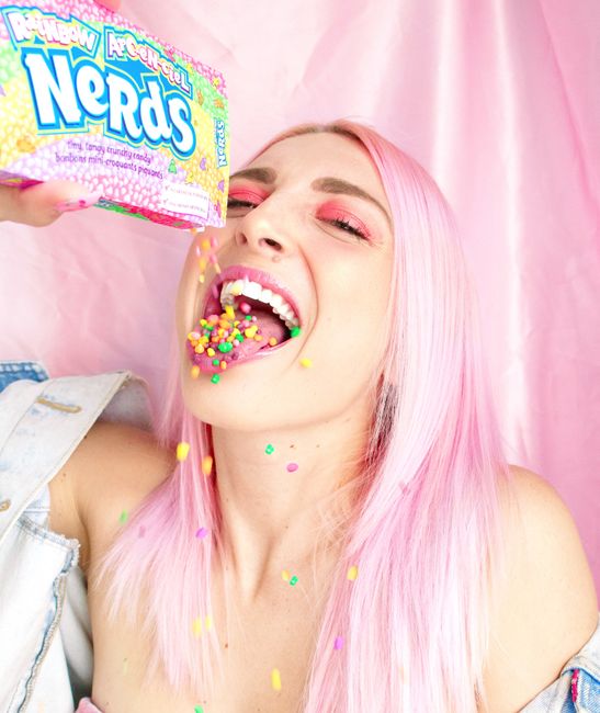 Young woman with pink hair playfully enjoying rainbow Nerds candy indoors.