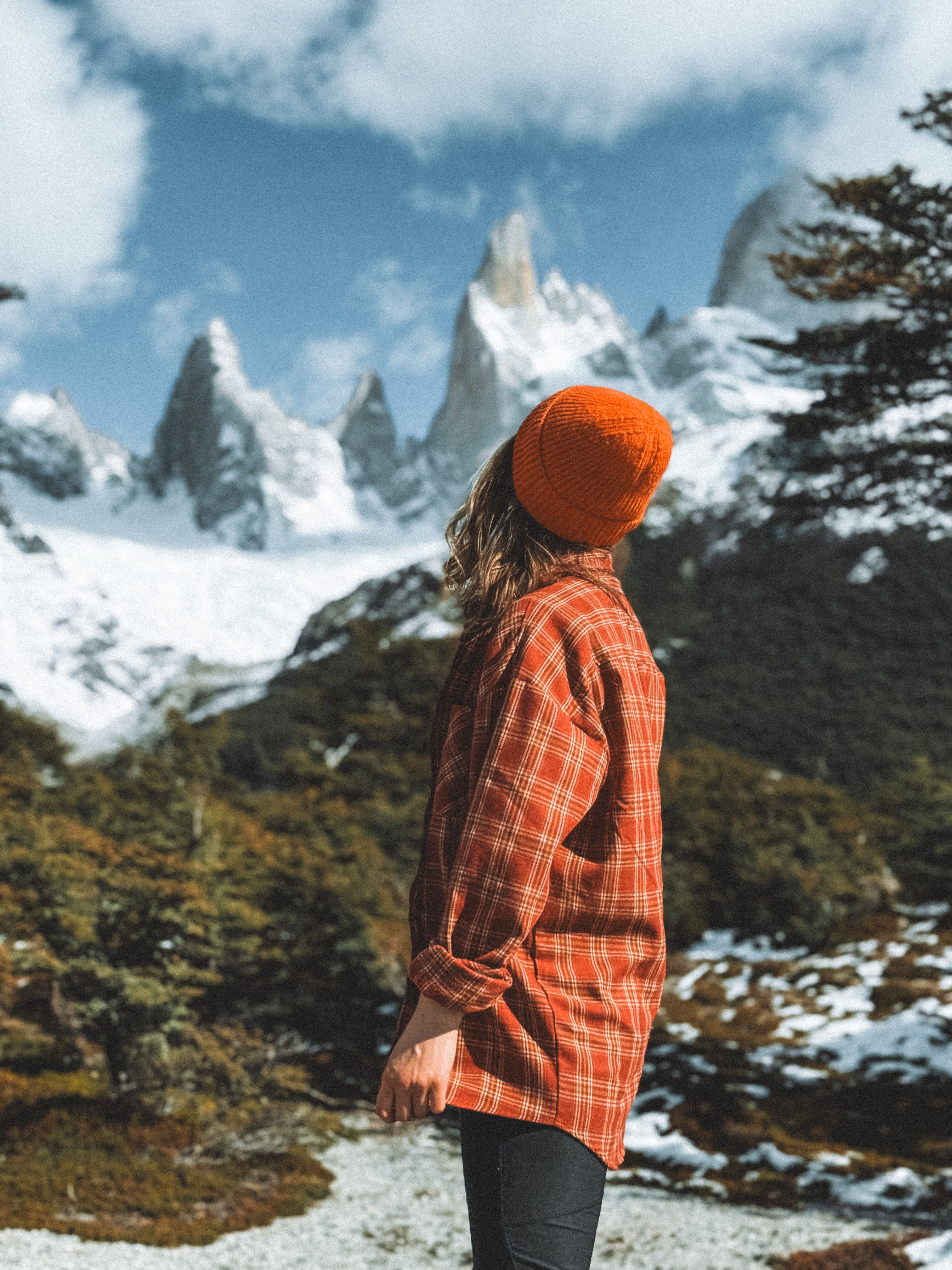 A woman stands gazing at the majestic Fitz Roy mountain in Patagonia, surrounded by snowy peaks and lush greenery.