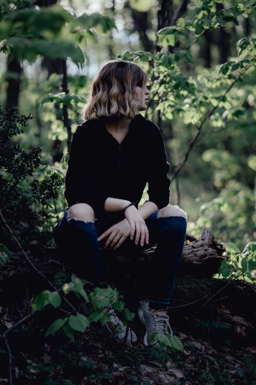 Free A woman sits alone on a log in a lush forest, surrounded by vibrant green foliage. Stock Photo