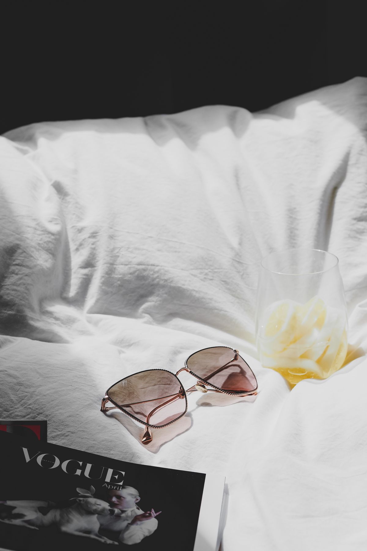 A pair of trendy sunglasses resting on a bed next to a Vogue magazine and a drink, in a Parisian setting. Stock Photo.