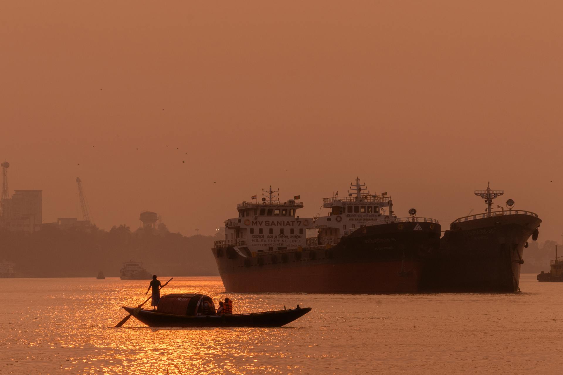 A serene sunset with a fishing boat and cargo ships silhouetted on the sea.