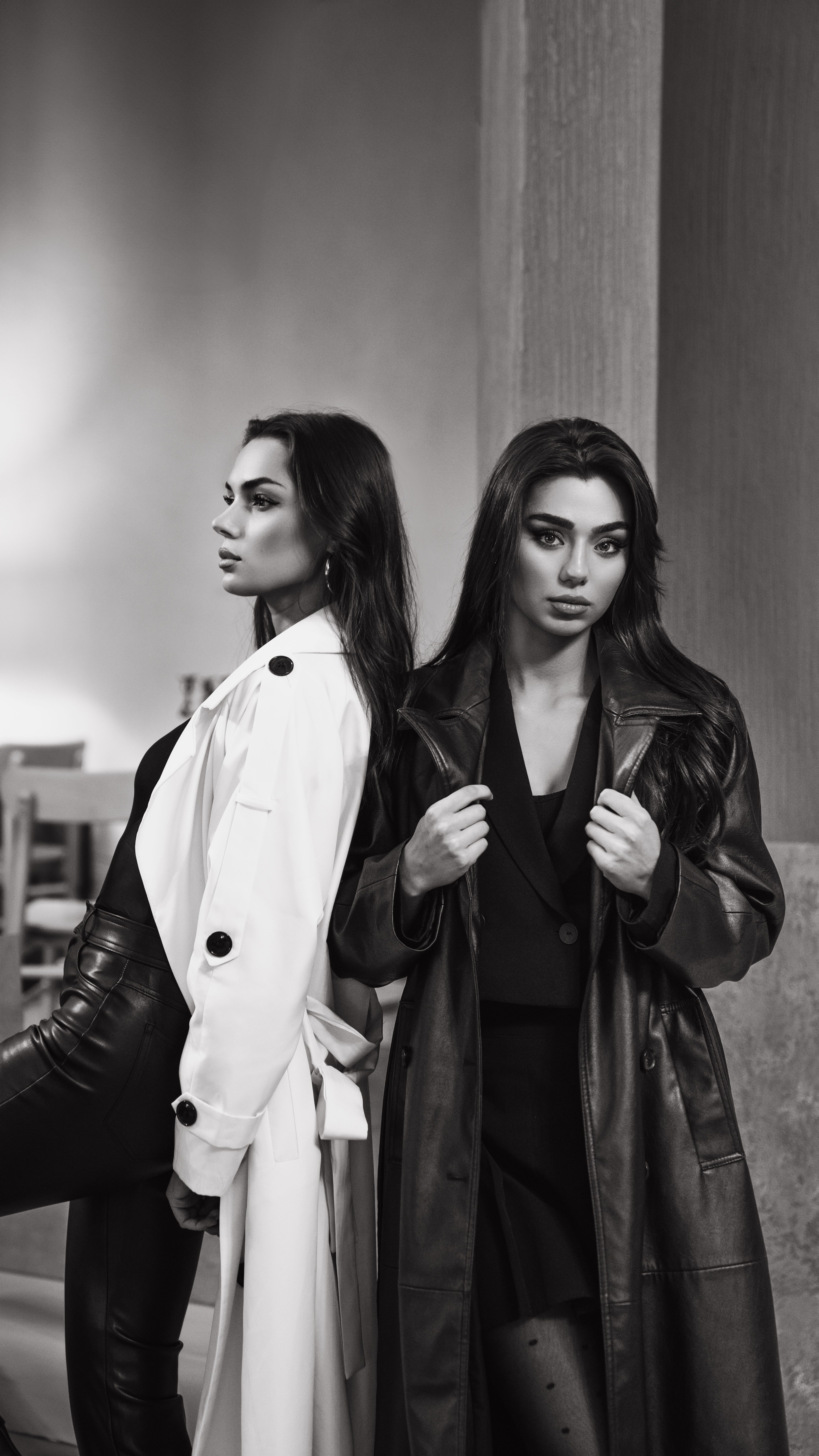 Two women in long black and white coats pose confidently in a stylish black and white portrait.