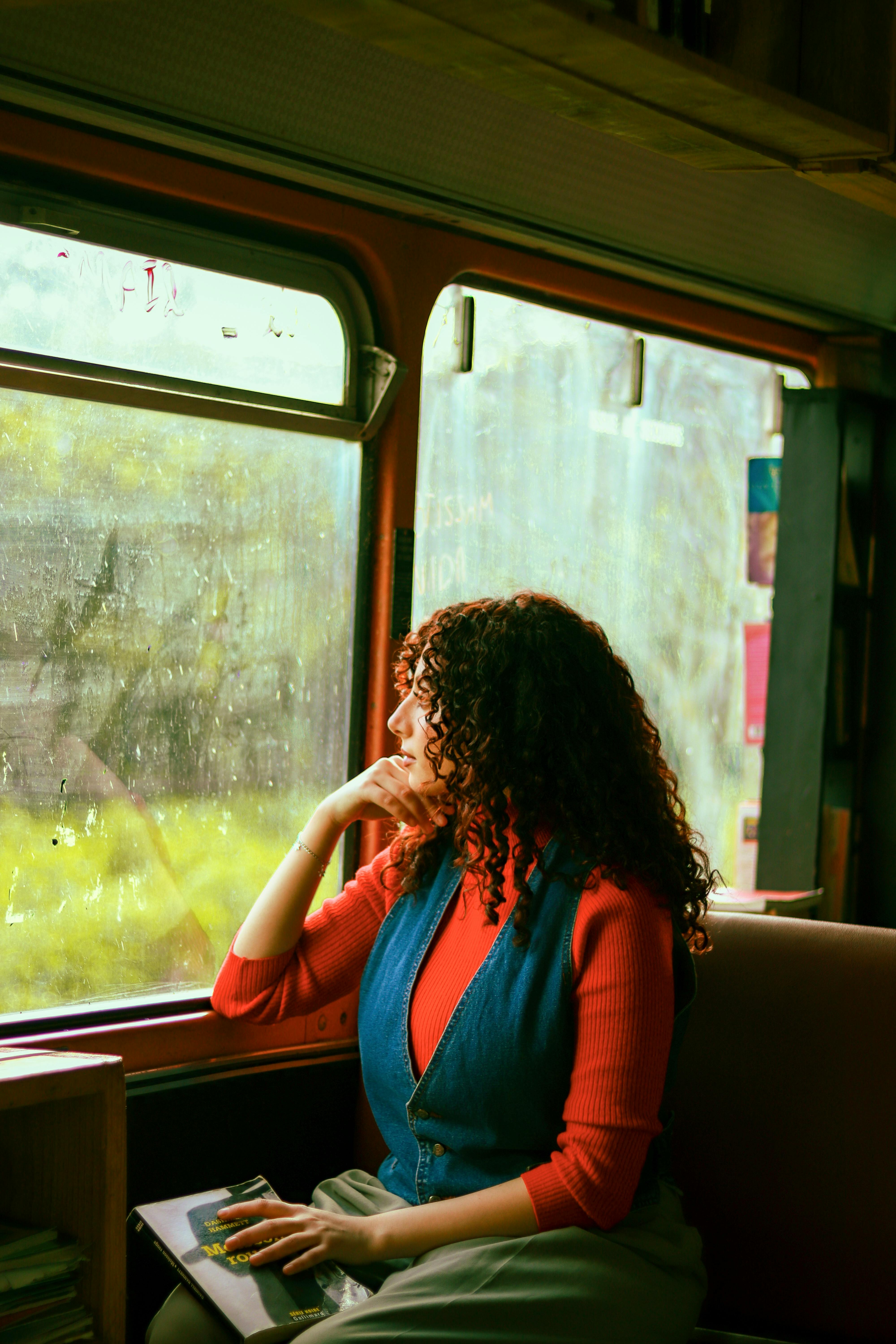 Curly-haired woman looks out train window while holding a book, during a daytime journey.