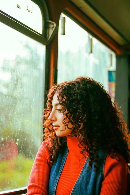 Free A young woman gazes out a bus window, wearing a red sweater and denim vest. Stock Photo