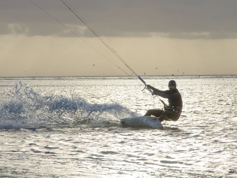 Kitesurfer in a wetsuit skimming across shimmering sea and kicking up spray