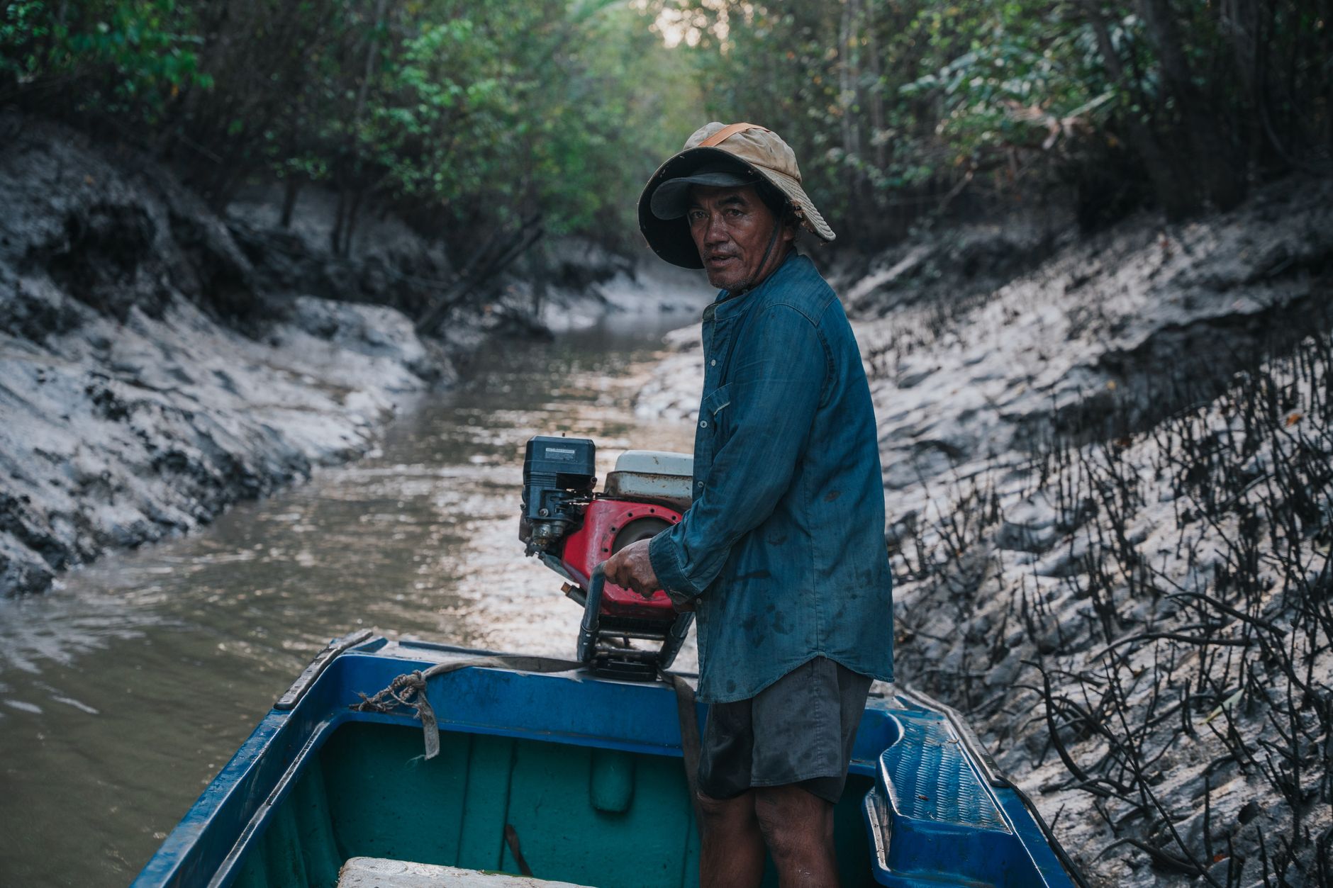 Fisherman operates motorboat through narrow canal surrounded by dense forest in Ho Chi Minh City, Vietnam.