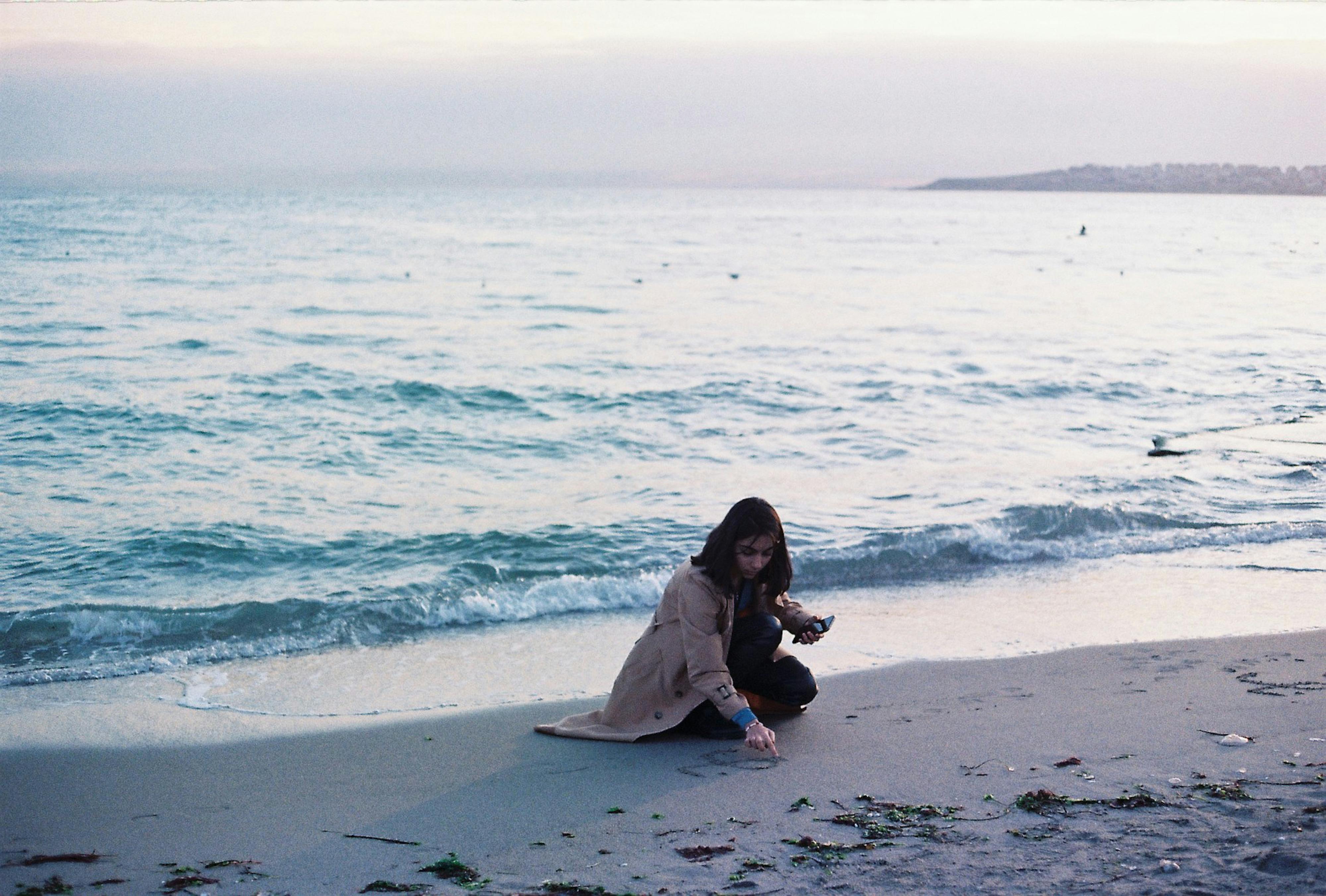 A woman squatting on a sandy beach drawing near the shoreline in the evening light.