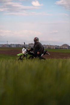 Man on a motorbike standing in a vast green field under a cloudy sky.