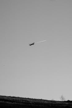 Black and white image of airplane flying over landscape with visible contrail in the sky.
