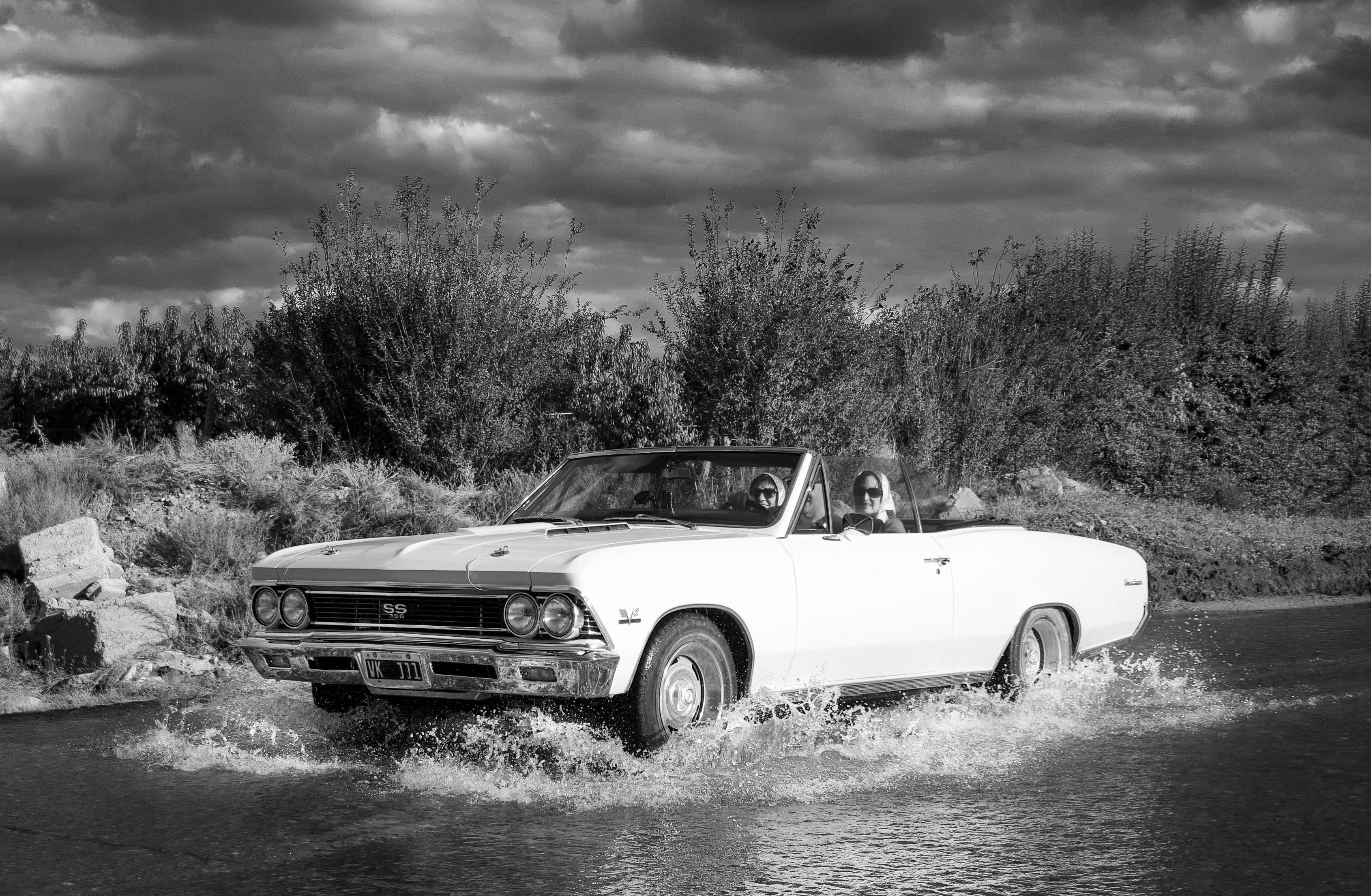 A vintage white convertible navigates through water with two adults onboard, captured in black and white. - Mendoza