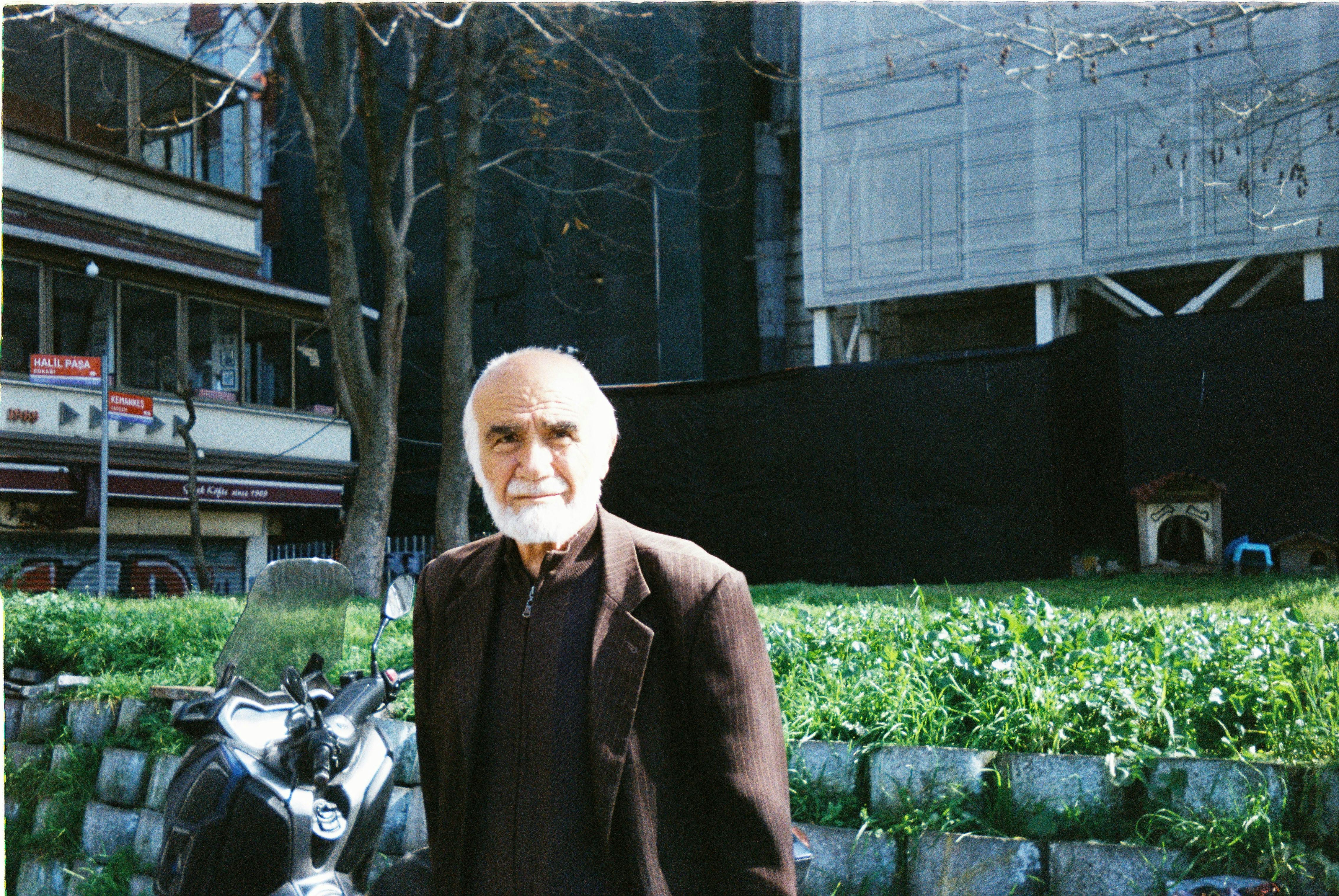 An elderly man with a beard and mustache in a brown jacket on a city street scene.