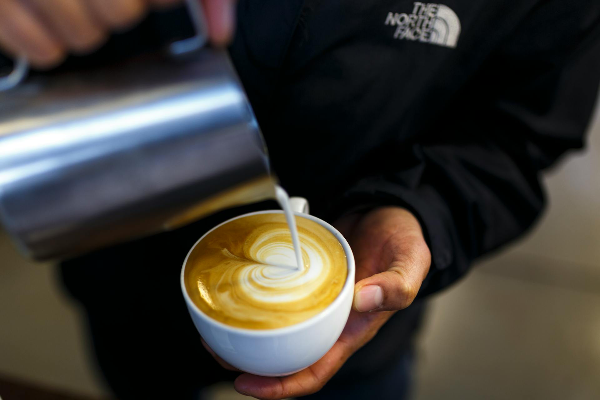 Close-up of barista pouring milk creating latte art in a cappuccino cup at a coffee shop.