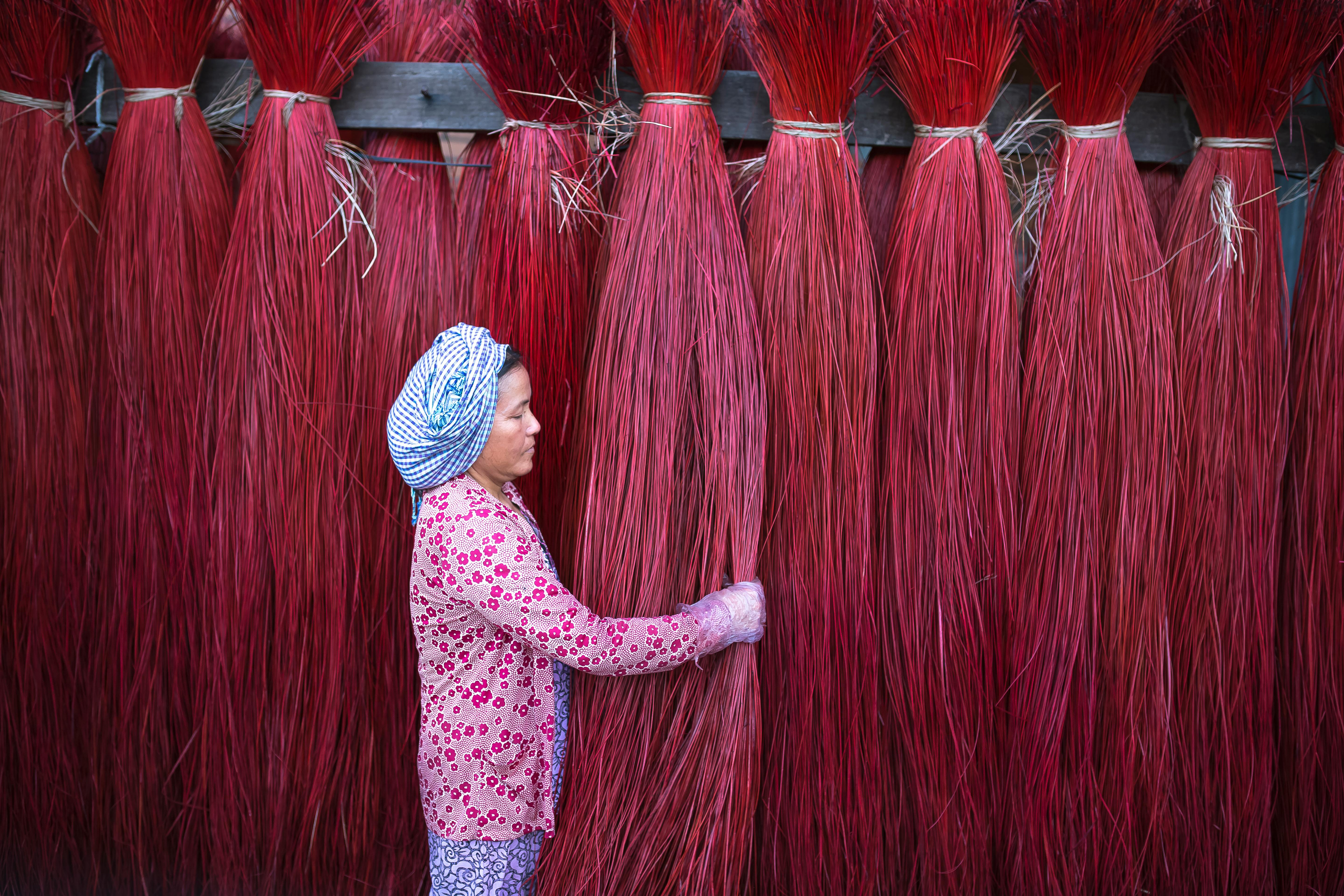 A woman in traditional attire handles colorful red straw bundles outdoors.
