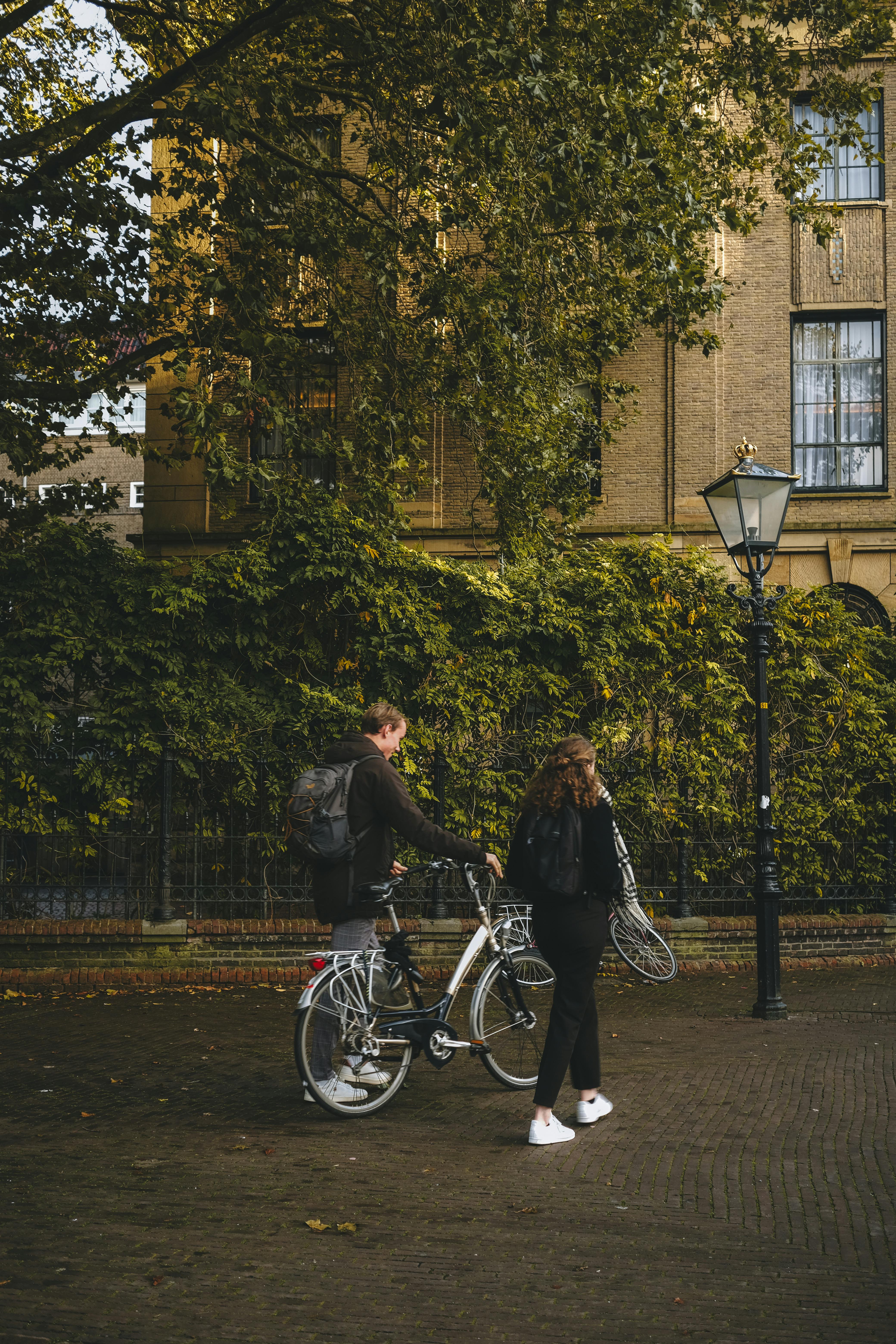 A couple strolls with bicycles on a charming cobblestone street lined with greenery and warm architecture.