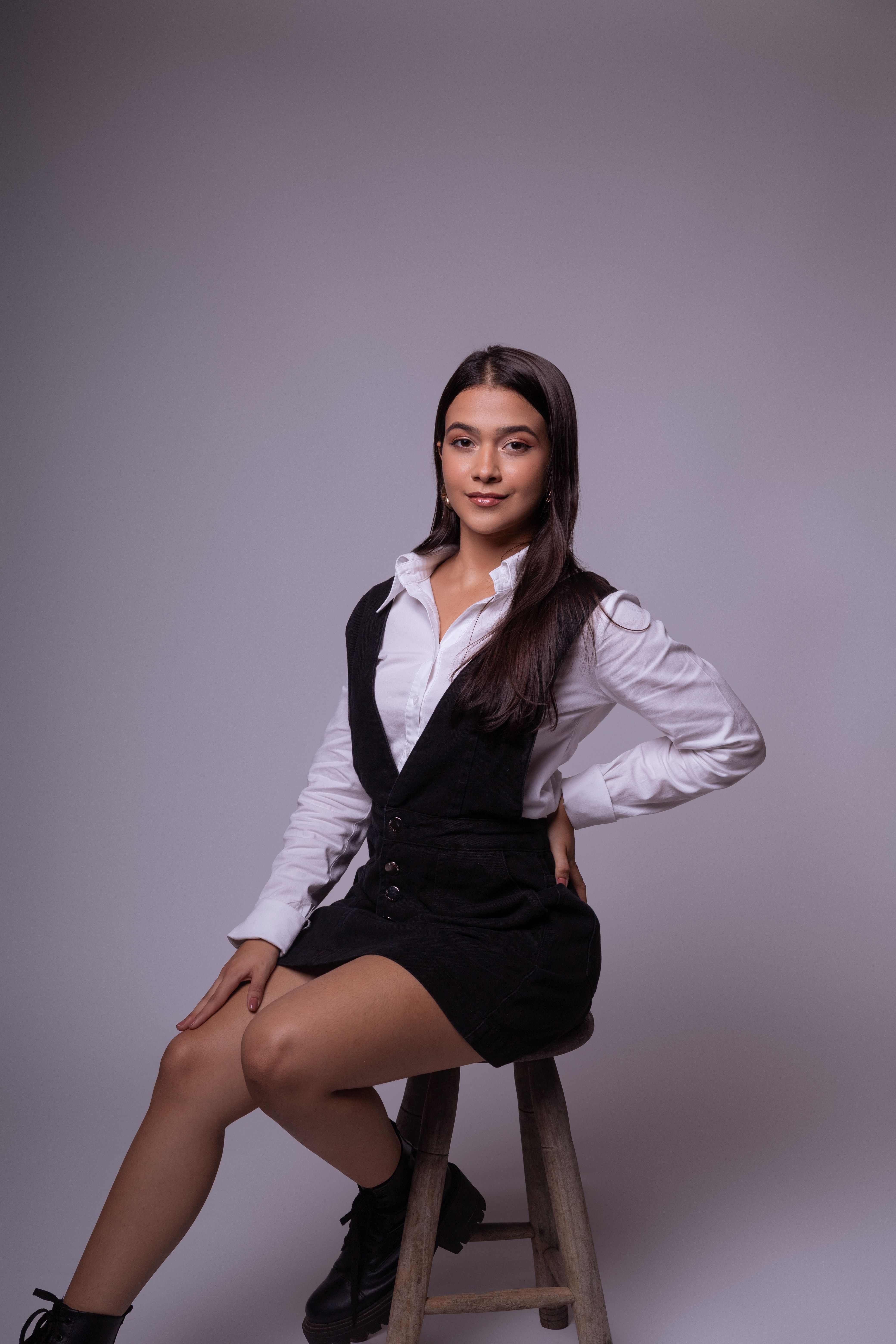 Young woman with long brunette hair seated on a stool, wearing a black overalls and white shirt against gray background.