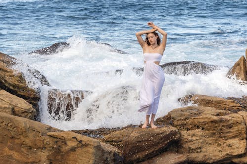 Free A woman in a white dress standing on rocks by the ocean Stock Photo