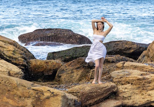 Free A woman in a white dress standing on rocks near the ocean Stock Photo