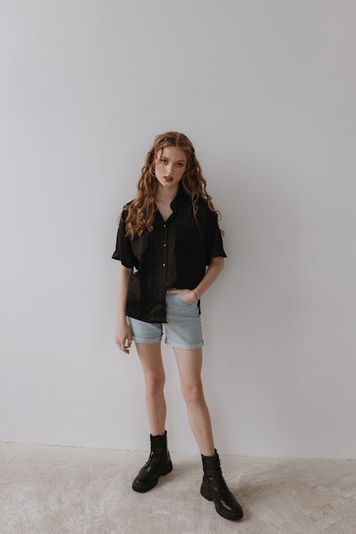 A woman in black shirt and denim shorts standing against a white wall