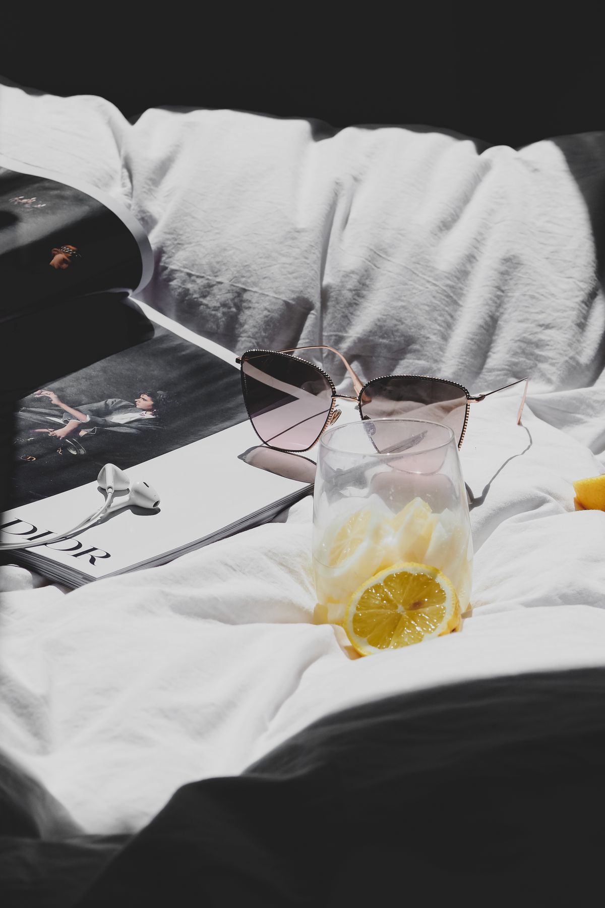 Stylish sunglasses on a bed with a fashion magazine and lemon water, evoking a relaxed Parisian vibe. Stock Photo.