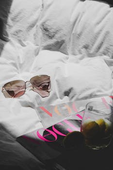 Chic setup with sunglasses, Vogue magazine, and a refreshing drink on a white bed, capturing a relaxed lifestyle in Paris.