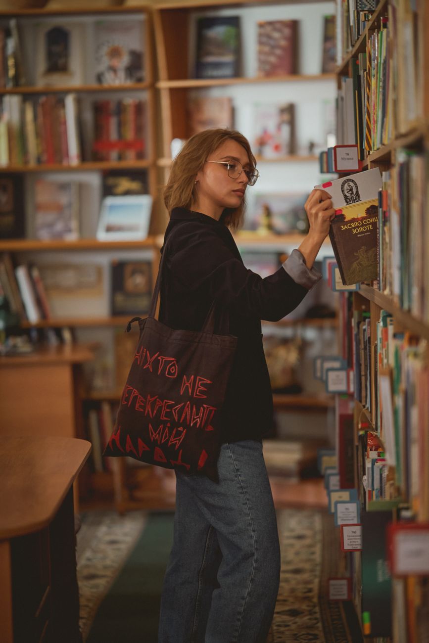 Reader reaching for a hardback while a tote hangs from her shoulder in a library aisle
