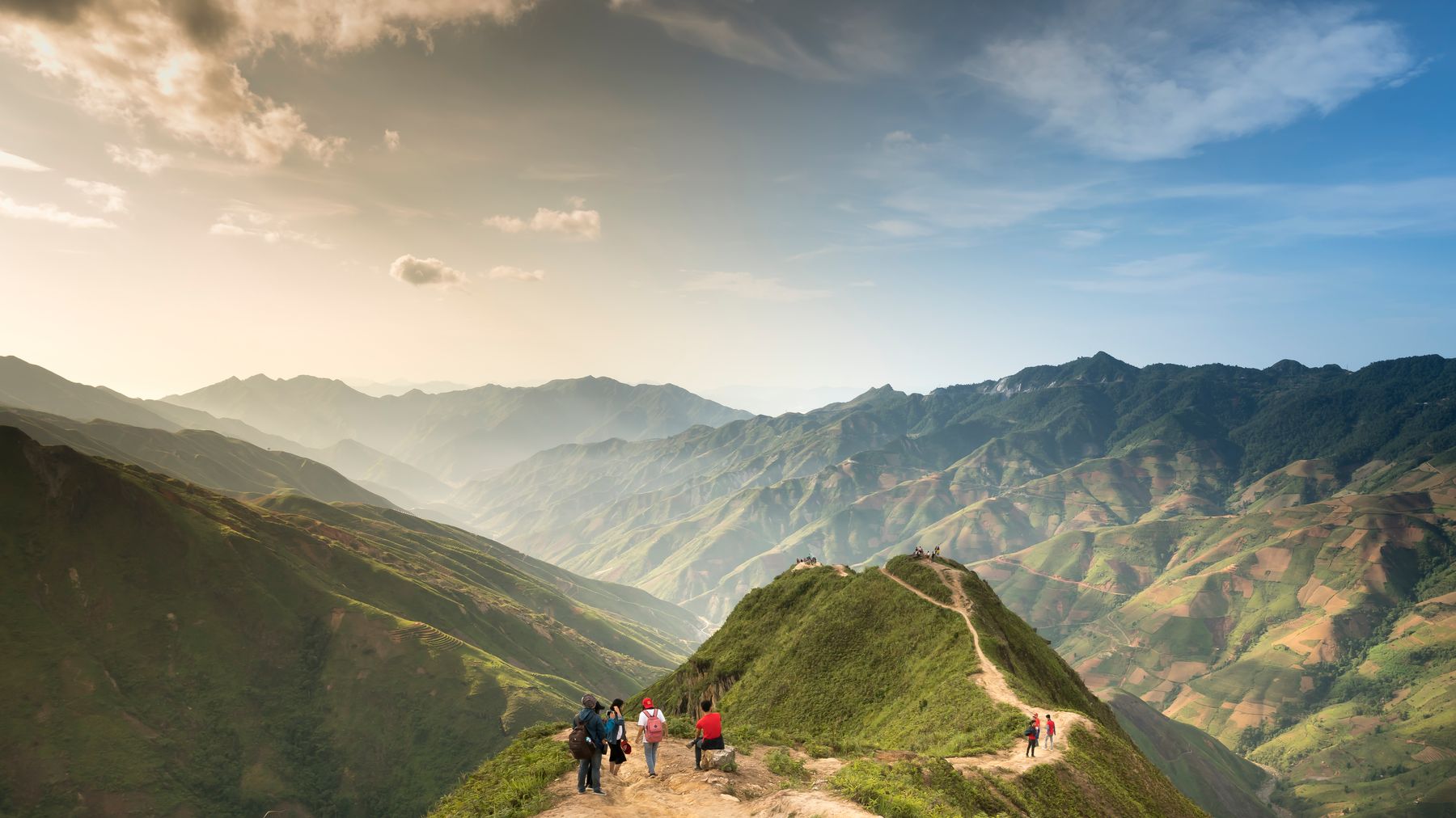Hikers walking along a scenic mountain trail with a stunning valley view under clear skies.