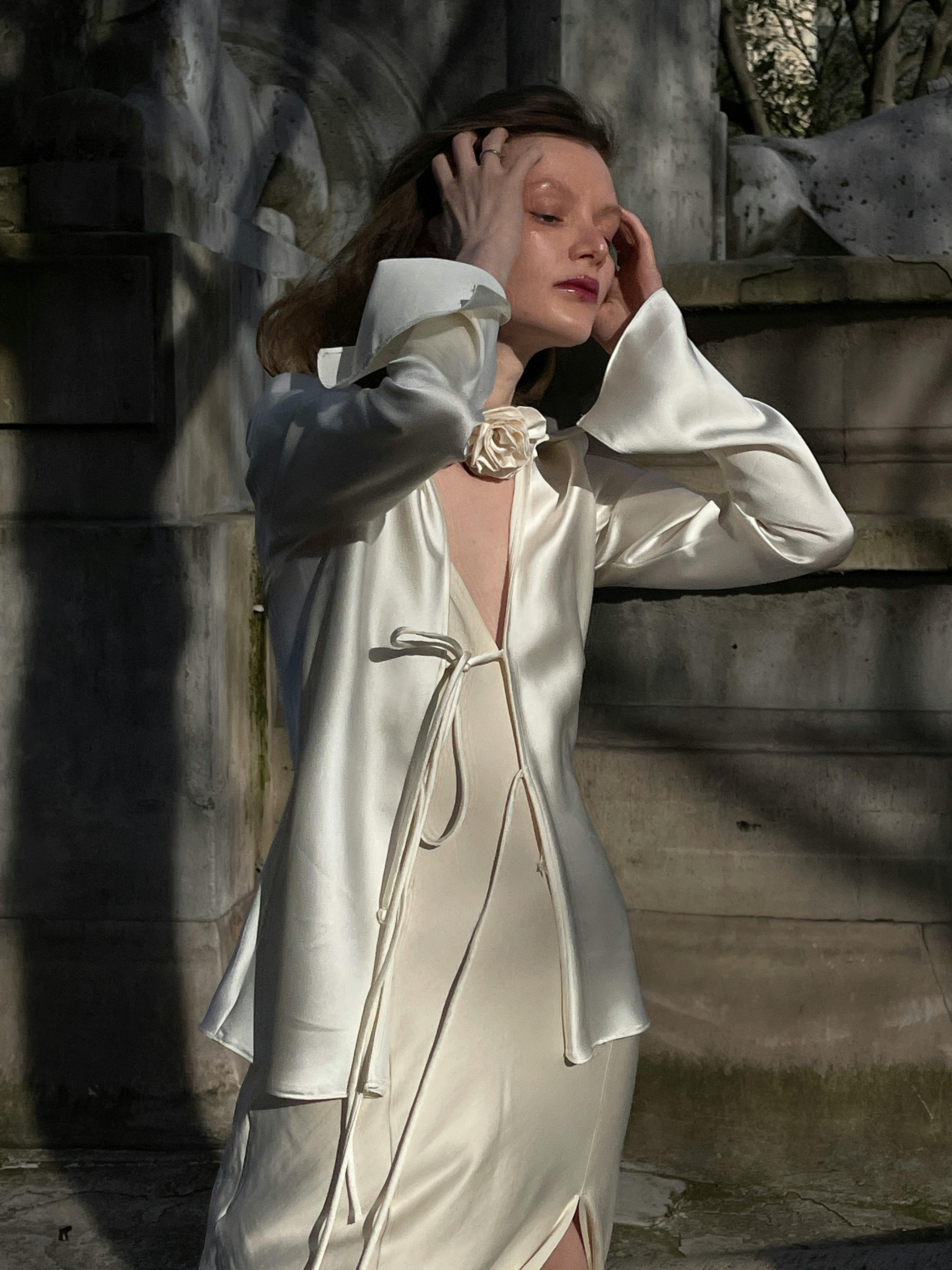 Woman in a delicate silk dress poses gracefully in sunlight with elegant architecture.