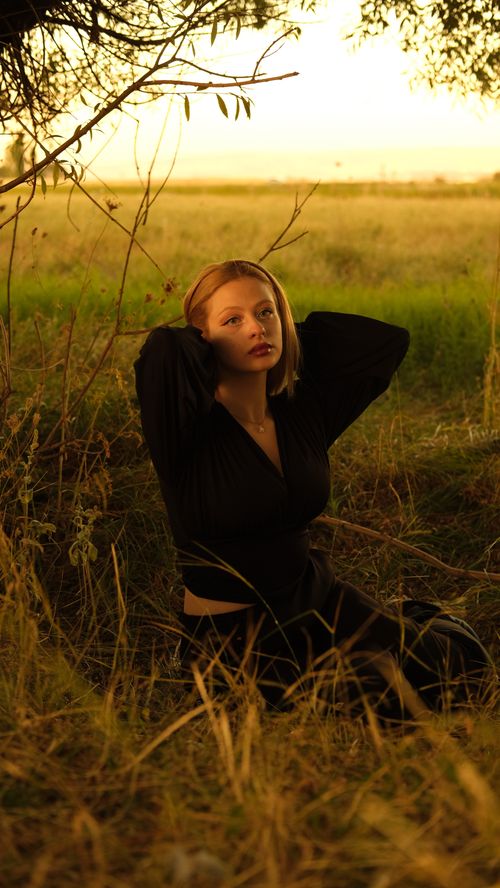 Free A woman in black fashion attire poses elegantly in a rural field during sunset, showcasing a serene and natural atmosphere. Stock Photo