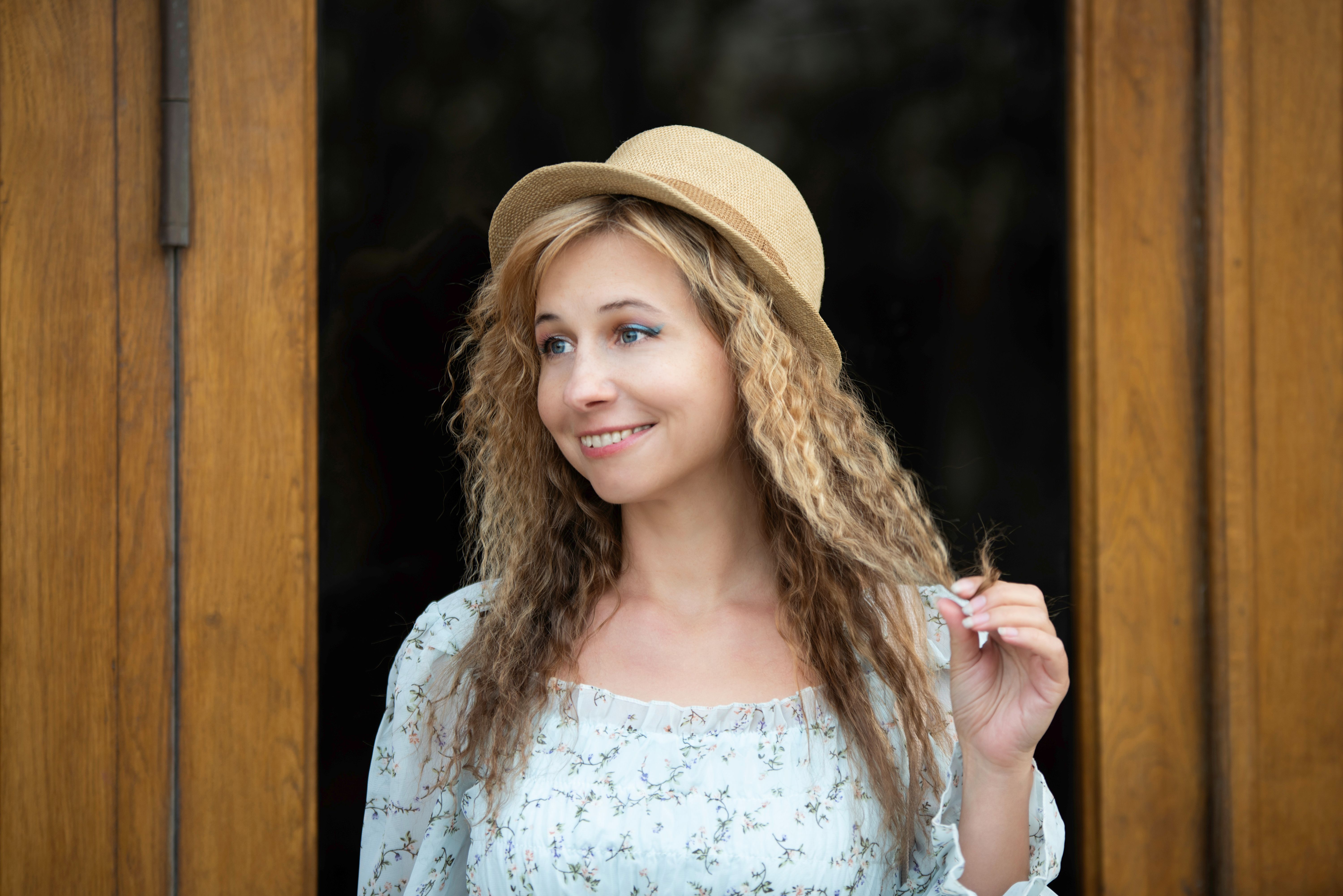 Elegant young woman in floral dress smiling and posing with a straw hat in front of a wooden door.