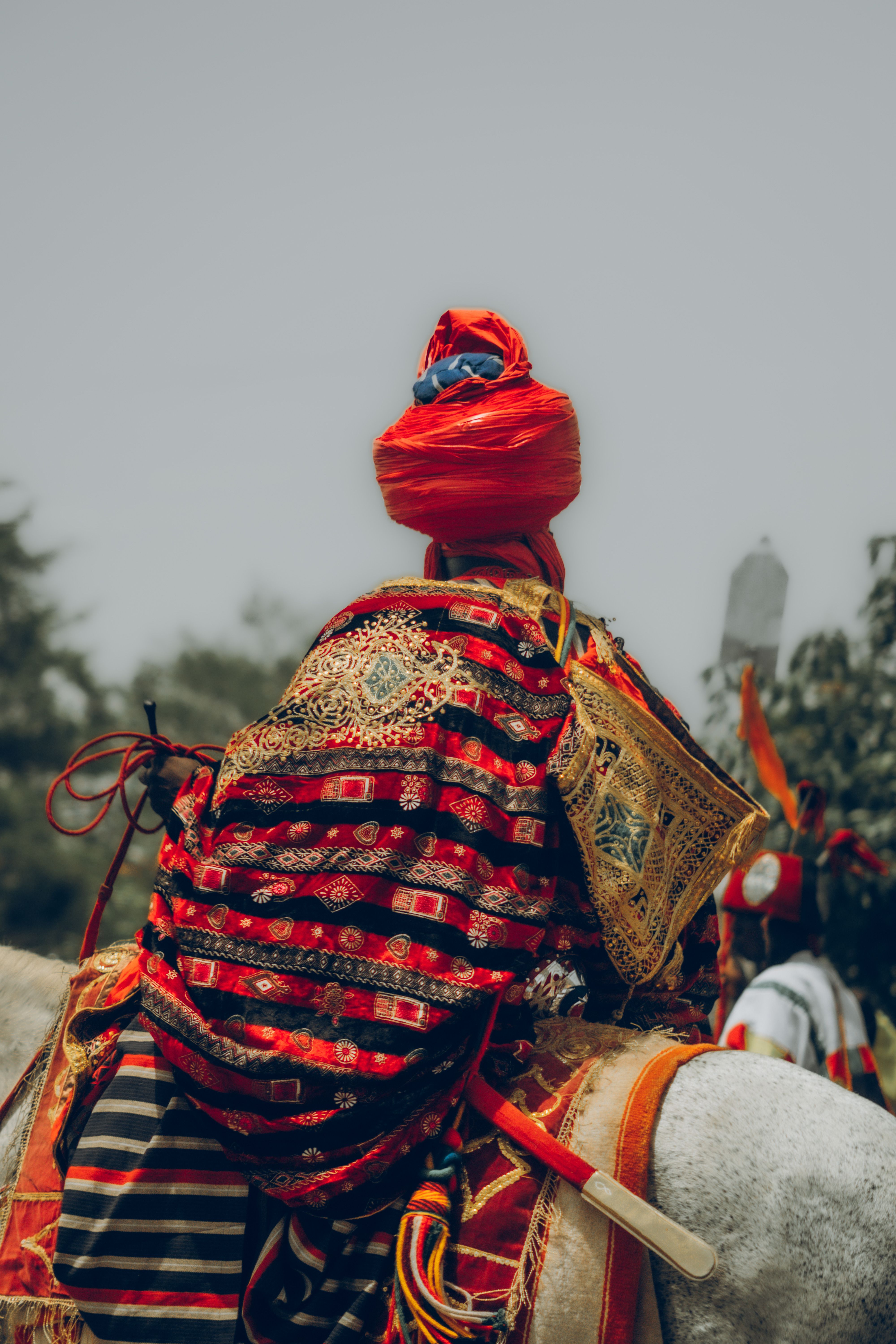 A man in traditional attire on horseback, adorned with bright textiles, set against an outdoor backdrop.