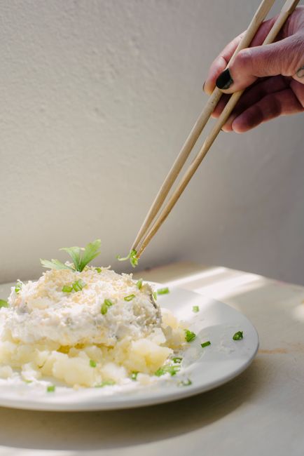 A hand using chopsticks to serve a delicious creamy potato salad garnished with herbs.