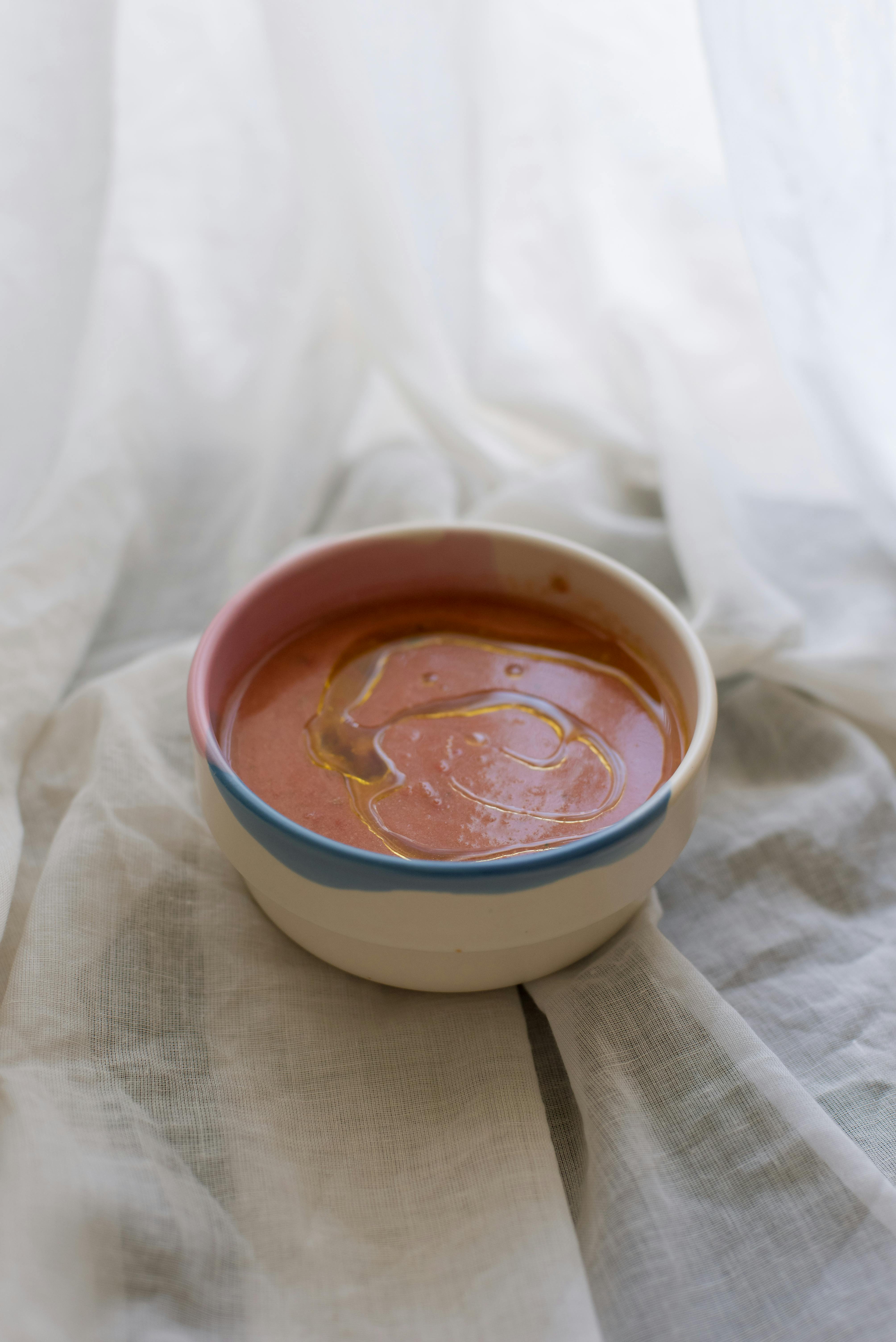 A rich tomato soup with olive oil drizzle served in a colorful ceramic bowl on a soft white fabric.