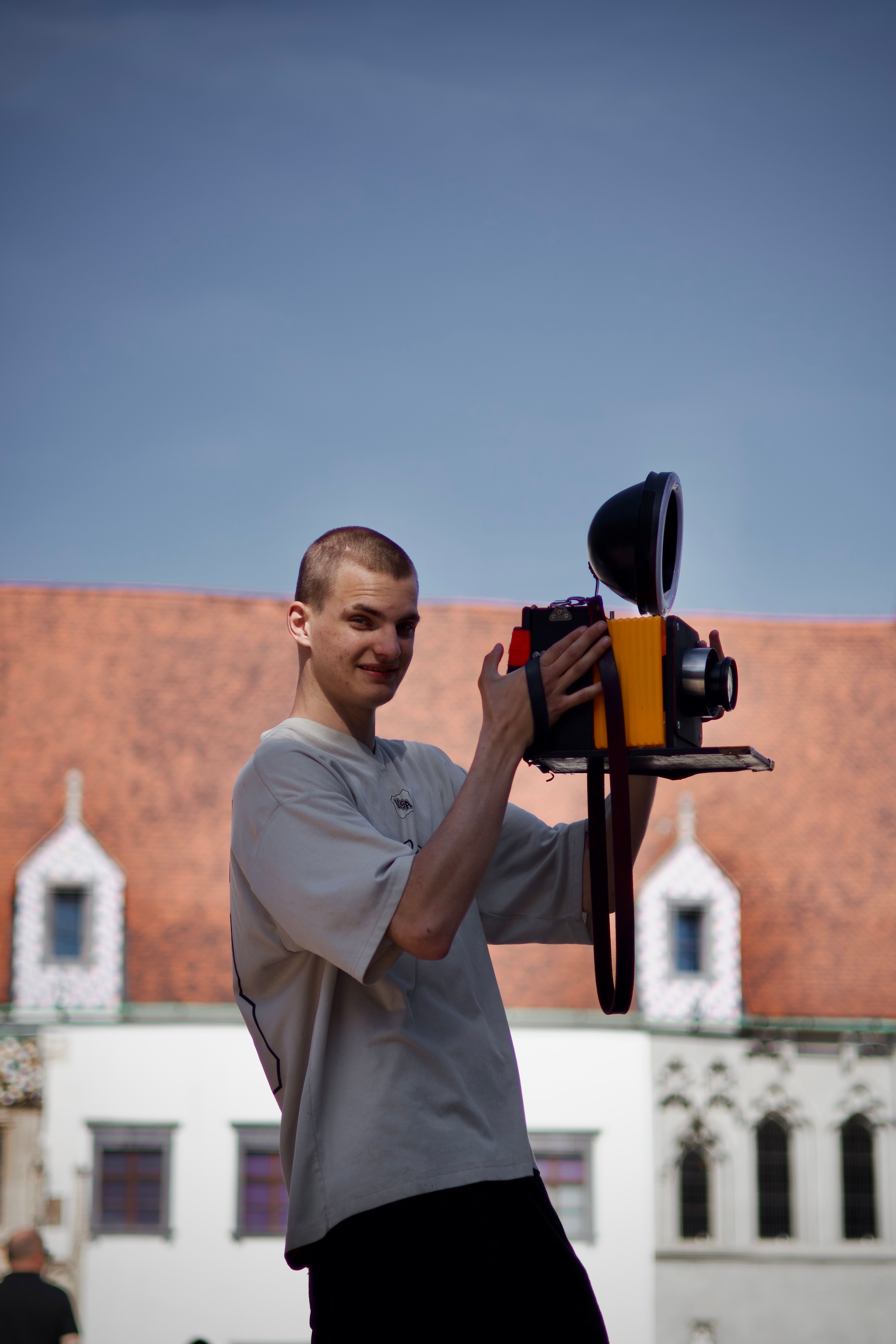 Young man holding a retro camera in front of a historic building on a sunny day.
