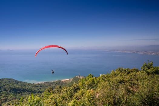 A paraglider flies over a beautiful coastal landscape with clear skies, offering a stunning view of the sea.