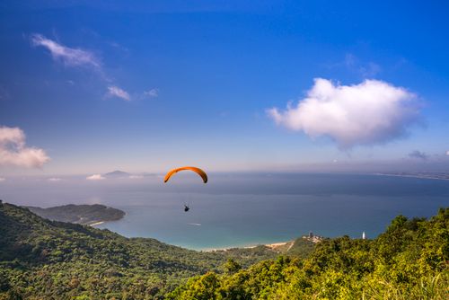 Free A paraglider soaring over a lush, mountainous coastal region with expansive ocean views and clear blue sky. Stock Photo