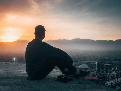 Free A person sits on a rooftop, silhouetted against a vibrant sunset sky, overlooking a cityscape. Stock Photo