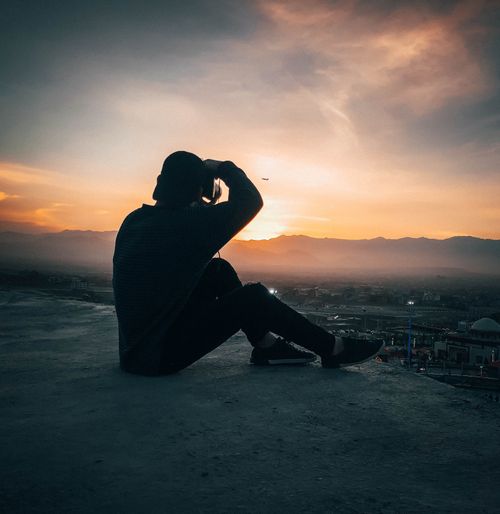 Free A man sits on a rooftop, silhouetted by a colorful and dramatic sunset horizon. Stock Photo