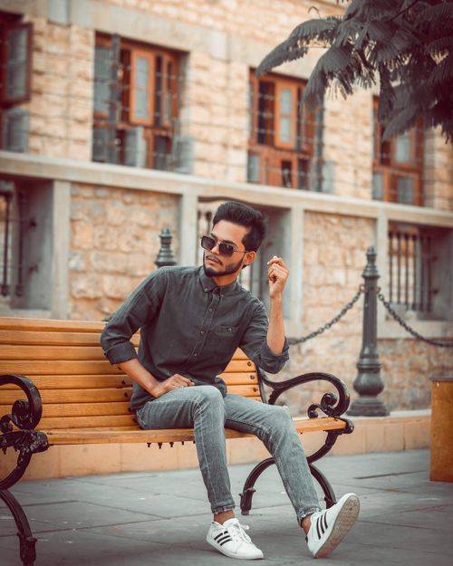 Free A fashionable young man sits on a wooden bench in an urban area, wearing sunglasses and a casual outfit. Stock Photo
