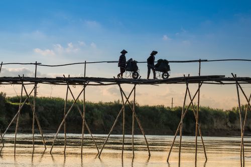 Free Two farmers push wheelbarrows across a rustic wooden bridge silhouetted against a scenic sunset sky. Stock Photo