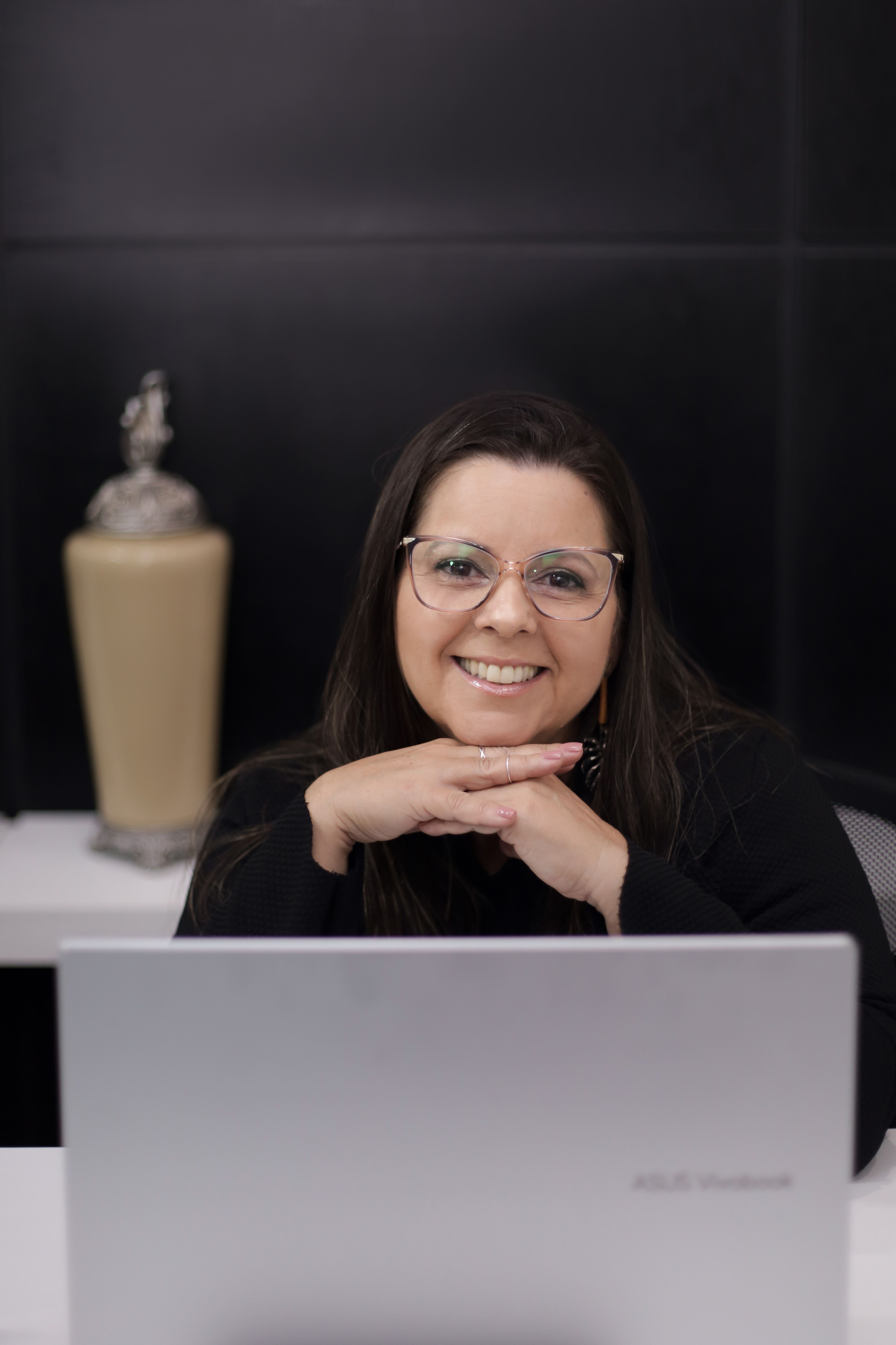 Smiling woman with glasses sits confidently behind a laptop in a sleek modern office setting.
