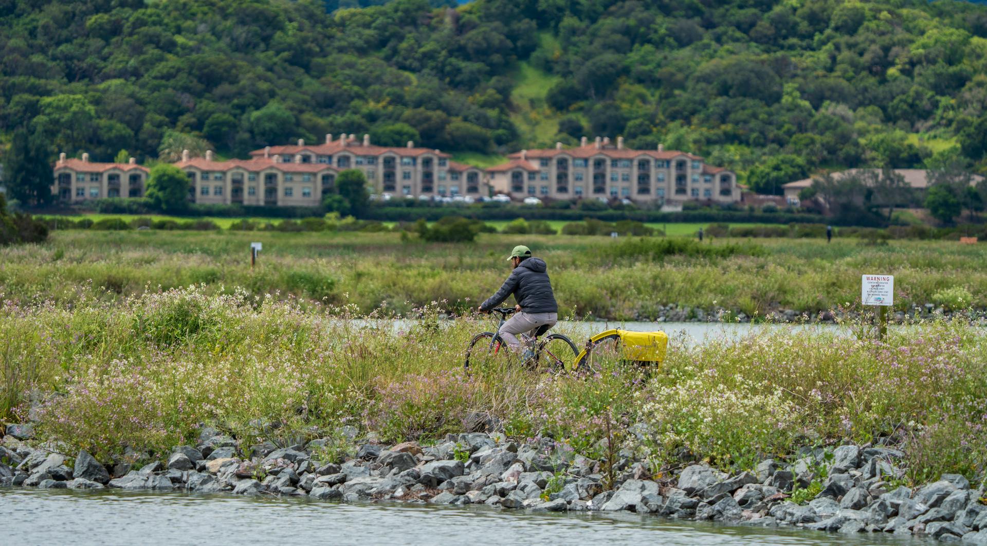 A person riding a bicycle with a trailer along a riverbank in a lush green landscape.