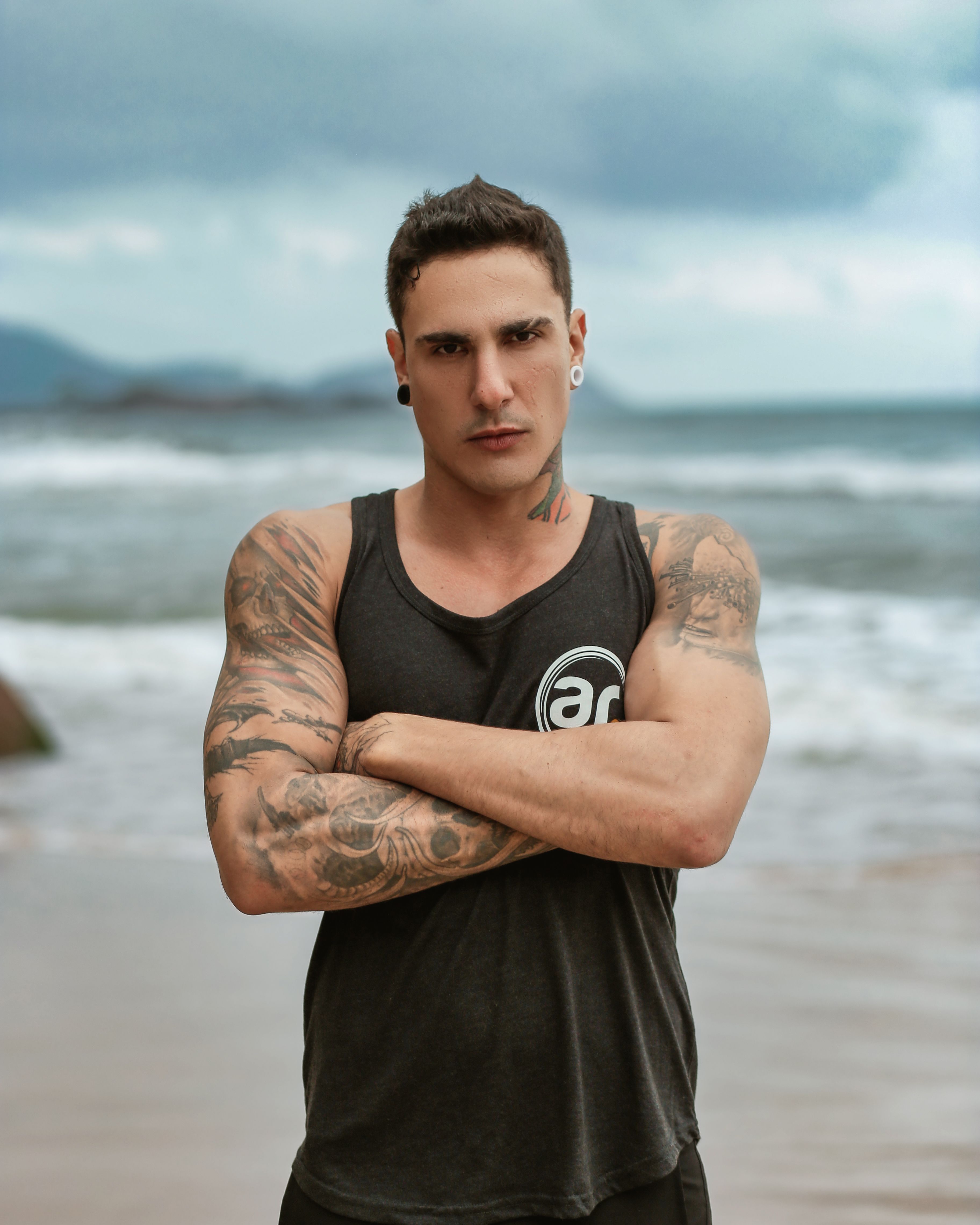 Portrait of a tattooed man with crossed arms on a beach with cloudy skies.