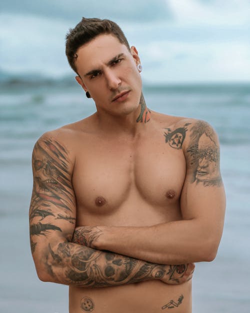 A man with tattoos on his chest and arms standing on the beach