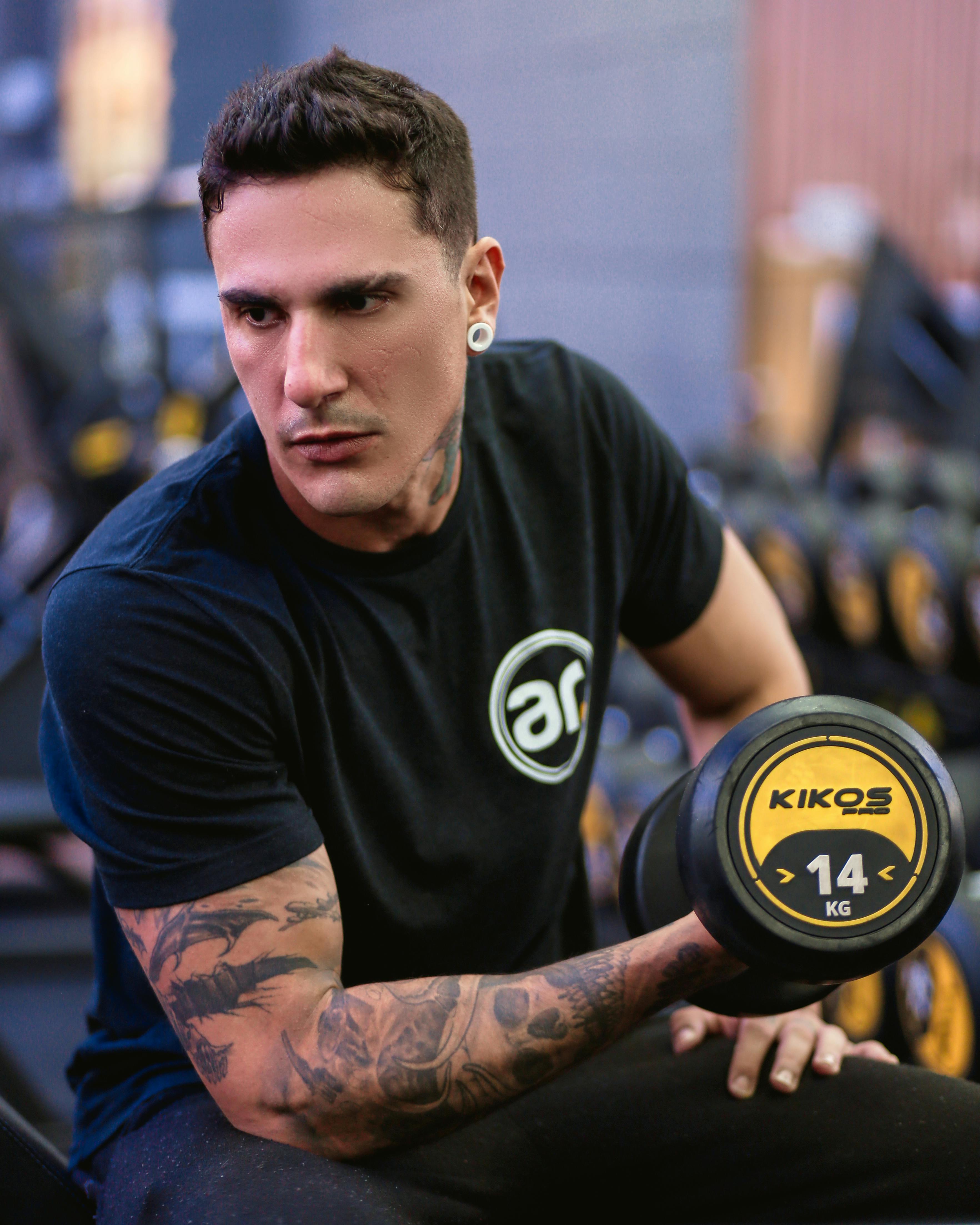 Portrait of a muscular man with tattoos lifting a 14kg dumbbell, focused on fitness in an indoor gym setting.
