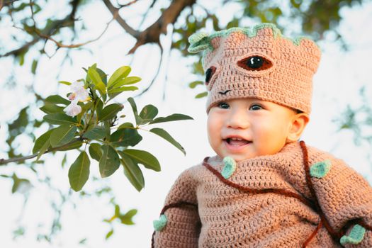 A smiling baby in a cute knitted costume is posed outdoors surrounded by branches and leaves.