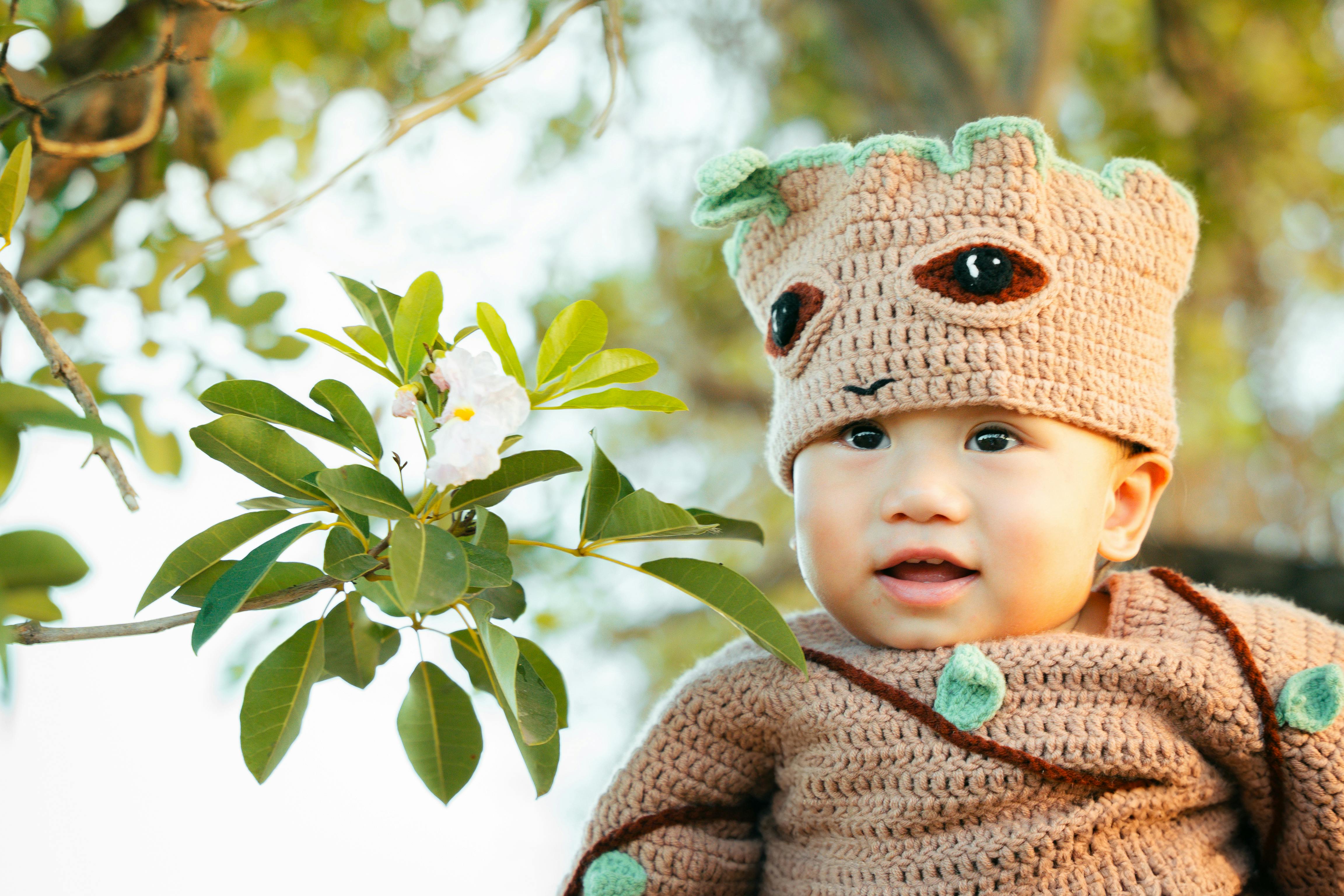 Cute baby in a knitted tree-themed costume smiling outdoors under green leaves.