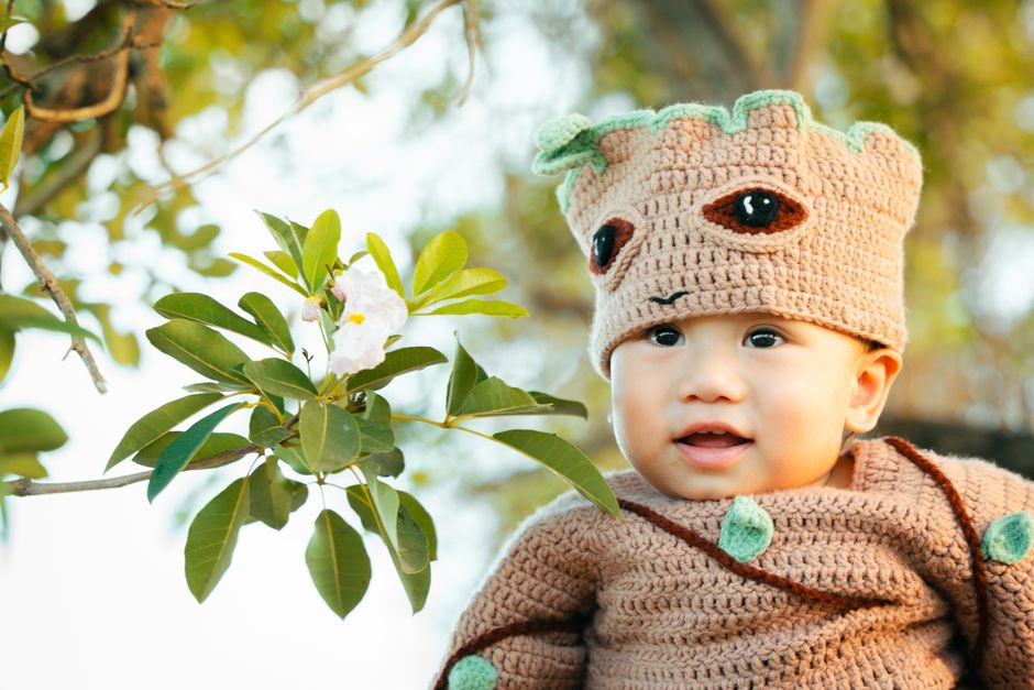 Cute baby in a knitted tree-themed costume smiling outdoors under green leaves.